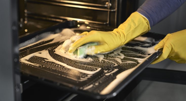 Person mit gelben Gummihandschuhen schrubbt ein schaumiges Backofenblech.