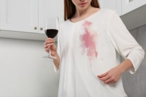 Frau mit Glas Rotwein betrachtet einen frischen Rotweinfleck auf ihrer weißen Bluse.