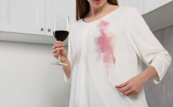 Frau mit Glas Rotwein betrachtet einen frischen Rotweinfleck auf ihrer weißen Bluse.