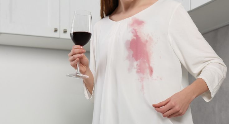 Frau mit Glas Rotwein betrachtet einen frischen Rotweinfleck auf ihrer weißen Bluse.