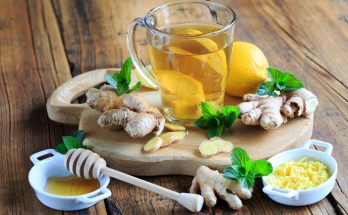 Glas Ingwertee mit Zitronenscheiben, frischem Ingwer, Minze und Honig auf einem Holzbrett.