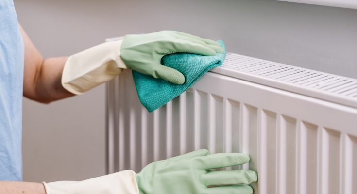 Person mit Gummihandschuhen wischt einen Heizkörper mit Tuch sauber.