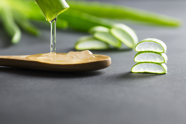 Transparentes Aloe-Vera-Gel tropft aus einem angeschnittenen Aloe-Vera-Blatt auf einen Holzlöffel neben mehreren frischen Blattstücken.