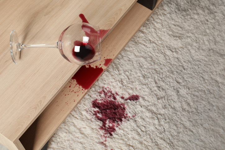 Verschütteter Rotwein aus einem umgekippten Weinglas mit Rotweinflecken auf einem hellen Teppich und einem Holztisch.