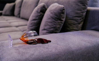 Umgekipptes Weinglas mit verschüttetem Rotwein und frischem Rotweinfleck auf einem grauen Stoffsofa.